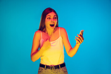 Pointing on herself, shocked. Caucasian young woman's portrait on blue background in neon light. Beautiful female model in casual. Concept of human emotions, facial expression, sales, ad. Copyspace.