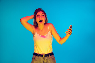 Holding phone, crazy happy winner, shocked. Caucasian woman's portrait on blue background in neon light. Beautiful model in casual. Concept of human emotions, facial expression, sales, ad. Copyspace.