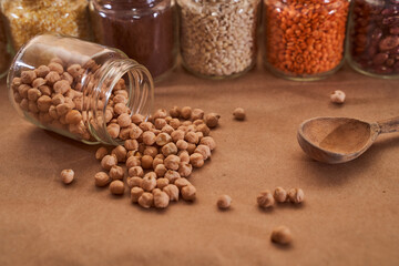 Legumes of chickpea scattered from a glass jar on against the background of glass jars with other superfood cereals d with a wooden spoon