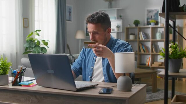 Cheerful young man makes an online purchase using a laptop and a bank card in his living room