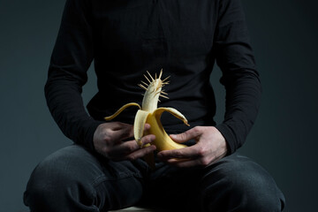 concept of genital cancer in men. A man in black holds a banana in his hands with thorns on his head, with a background on the male penis. On a gray background.