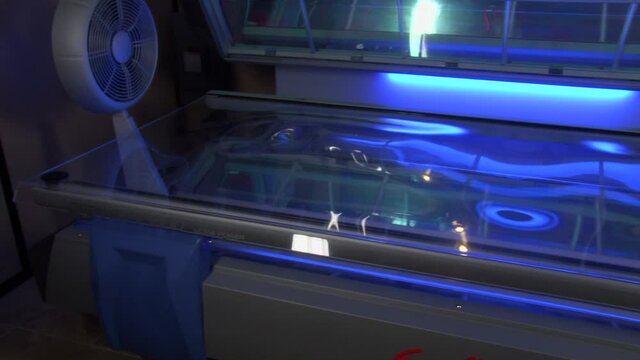 Blue sunbed with lid open showing clear bed surface, camera panning left