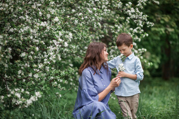 Fototapeta premium Happy and beautiful mother and son in clothes of blue tones are hugging next to a blossoming apple tree. The concept of spring, motherhood, happy family, relationships.