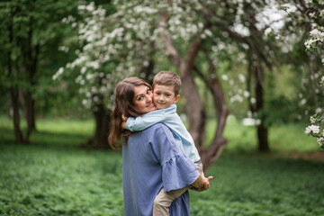 Happy and beautiful mother and son in clothes of blue tones are hugging for a walk in a blossoming apple orchard. The concept of spring, motherhood, happy family, relationships.