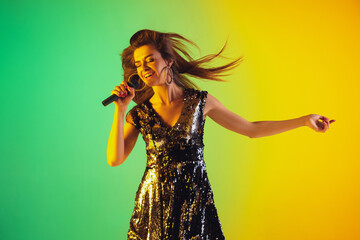 Caucasian female singer portrait isolated on gradient studio background in neon light. Beautiful female model in dress with microphone. Concept of human emotions, facial expression, ad, music, art.