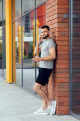 Portrait of handsome young man listening to music after running. Side view of young man in sportswear using his smart phone. Athlete man in earphones is having rest and listening to music. Brick wall.