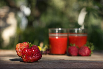 Large ripe strawberries on a background of two glasses with strawberry smoothie, a wooden table on a background of green garden. Tasty and healthy berries