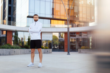 Fototapeta na wymiar Cheerful male athlete resting after street workout session. Stylish urban young man in sportwear with a stylish hairstyle and beard stands near modern buildings in the city. Urban environment.