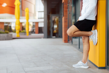 Close up legs of man standing close to a yellow wall with gray sport shoes. Modern sporty man making pause and stretching after jogging / exercise in urban environment.