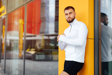 Young man standing in front of a building. Portrait of handsome young bearded man standing against yellow wall with arms crossed. Fashion style portrait of a muscular, handsome guy in city environment