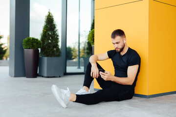 Man Sitting Listening Music Earphones  Having Rest Concept. Handsome jogger sitting in urban area listening to music looking at the phone.Young male athlete training and doing workout outdoors in city
