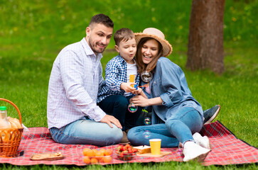 Weekend family pastimes. Happy boy blowing soap bubbles with his mom and dad on picnic outdoors