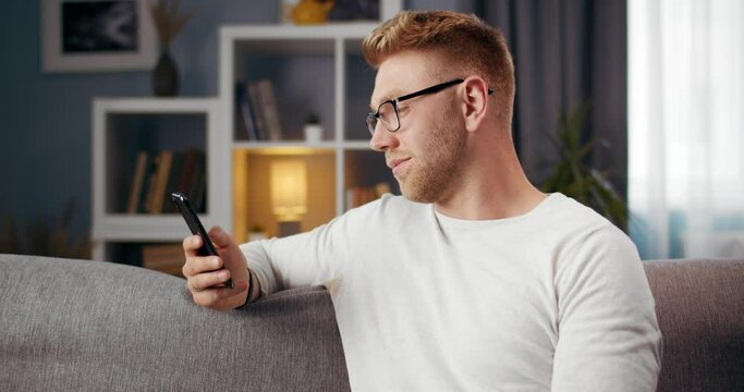Happy bearded guy in glasses relaxing on grey couch and looking at smartphone screen. Young man with stylish haircut using mobile during free time at own apartment.