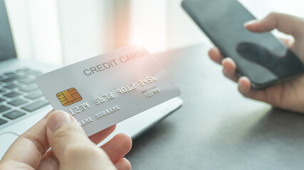 Woman hands holding a white credit card and using a smartphone for shopping online or internet banking with laptop background. Online shopping concept.