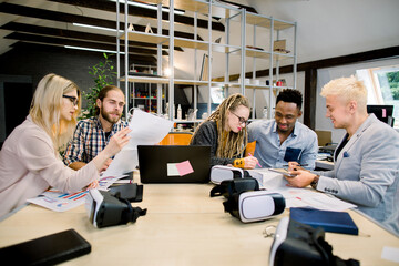 Business meeting. Creative team of young multiethnic colleagues, students, IT developers brainstorming while sitting at the office table together and working with laptop, tablet and papers