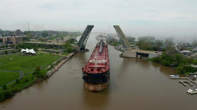 Aerial pull out rising shot of cargo ship passing under open lift bridge in Lorain Ohio with fog over Lake Erie in background