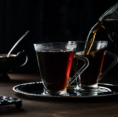 A close up of two cups of tea on a metal tray with tea being poured into one, against a black background.