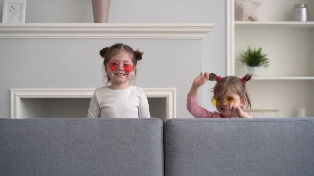 Funny little girls play at home, jump out from sofa, scare, scream and laugh, look at camera. Happy family concept.