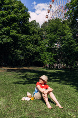 A redheaded boy sits in a park with street food and drink, sunny day.