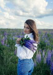 Beautiful brown-haired girl in a lupine field, lupine flowers. A girl with dark skin, dressed in jeans, a white blouse and a white hat, sits in a field of lupins.