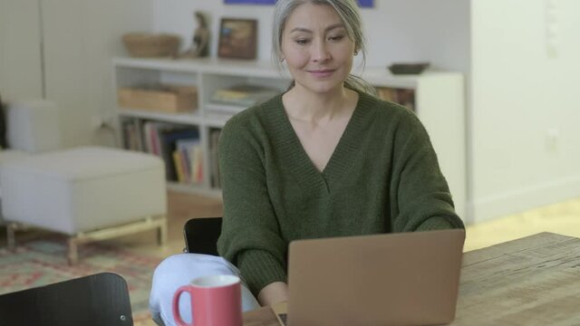 Smiling attractive mature woman with long gray hair using laptop computer while sitting by the table indoors