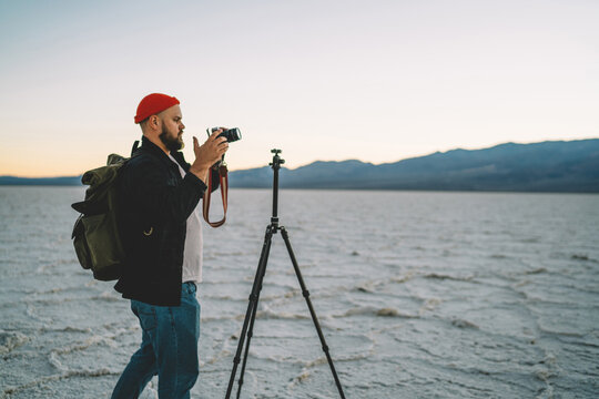 Skilled male photographer using modern camera and tripod during photo session in death valley, professional journalist taking picture of wild nature during expedition in Badwater basin in evening.