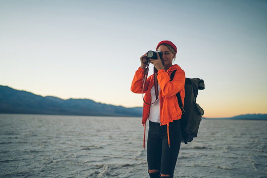 Skilled female photographer in trendy orange jacket making picture of sunset in desert during journey, young woman taking photos in twilight on professional camera exploring death valley on vacation