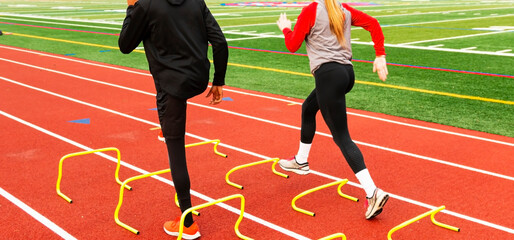 Rear view of wo athletes running over yellow mini hurdles on a track