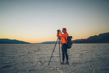 Skilled female photographer with equipment taking photos on camera and tripod during evening in desert environment, young woman wanderlust shooting video of sunset  in expedition in Badwater .