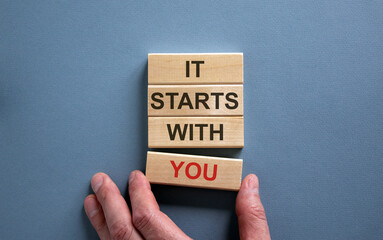 Wooden blocks form the words 'it starts with you' on blue background. Male hand. Business concept. Copy space.