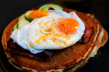 Three pancakes with fried eggs, avocado slices, grilled bacon and melted cheddar all in one black ceramic plate ready to be eaten