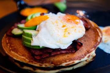 Three pancakes with fried eggs, avocado slices, grilled bacon and melted cheddar all in one black ceramic plate ready to be eaten