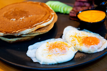 Three pancakes with fried eggs, avocado slices, grilled bacon and melted cheddar all in one black ceramic plate ready to be eaten