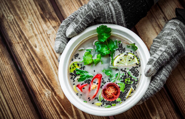 Woman hands in gloves holding plastic bowl of vegetable soup on wooden table background