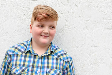 Plump red-haired boy is smiling. A boy in a plaid shirt stands on a wall background and smiles.