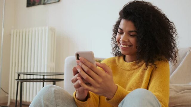 Smiling african attractive woman using smartphone while sitting on the floor in living room