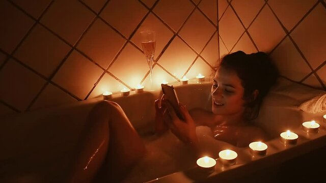 tanned sexy young girl lies in a foam bath with a smartphone and looks with a smile at the phone screen flipping through the tape