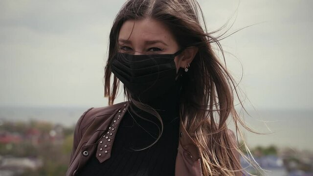 Brown-eyed young girl in a black protective antiviral mask. Hair blows in the wind in slow motion. Portrait of a girl during an epidemic Covid