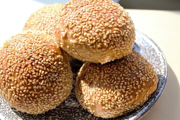 Homemade rye buns with sesame seeds stacked on top of each other on a ceramic plate. In the bright rays of the day sun. Natural shadows