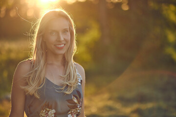 Smiling attractive blond woman backlit at sunset by the warm glow of the sun through leafy green trees in a blurred rural background with copy space
