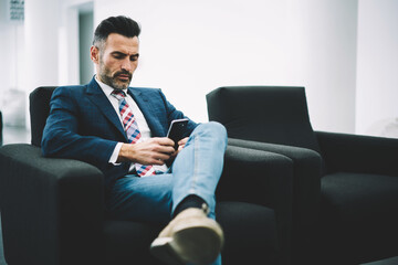 Pensive mature entrepreneur dressed in formal wear reading financial news on website on smartphone via 4G internet sitting on armchair in office interior.Concentrated proud ceo checking mail