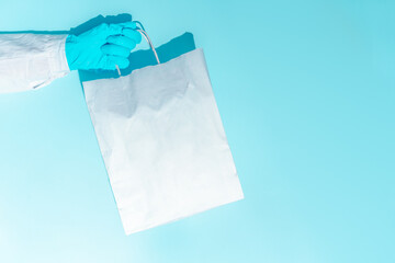 hand in gloves holding shopping paper bag. Concept of consumerism, shopping symbol during pandemic time. Copy space trend pastel color. Hard shadows, morning light.
