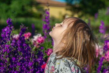 Naklejka premium A little girl in the garden with closed eyes inhales the aroma of tall bright flowering peonies and larkspur. The scent of flowers, pleasure, enjoying.