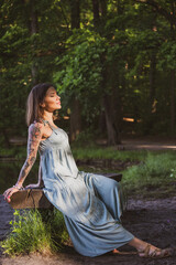 beautiful natural woman with tattoos sitting on a bench in a forest on the shore of the lake, illuminated by the morning sun