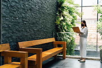 Asian businesswoman working on laptop while standing outside office building. She wears a face mask during covid 19 pandemic. - Full length