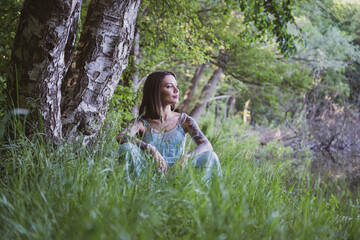 Fototapeta na wymiar beautiful natural woman with tattoos sitting on the grass, leaning on her knees on the shore of a forest lake, wearing a blue dress and looking at the water