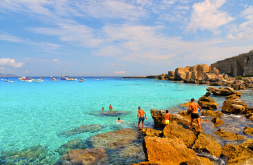 paradise clear torquoise blue water with boats and cloudy blue sky in background in Favignana island, Cala Rossa Beach, Sicily South Italy.