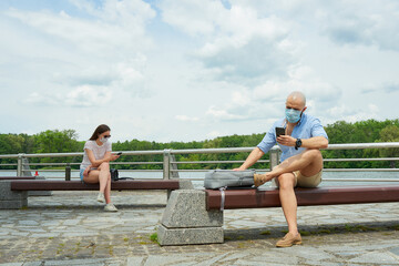 A bald man and a woman in medical face masks using smartphones keeping social distance sitting on different benches on a promenade. A guy and a girl resting in social distancing in the park.