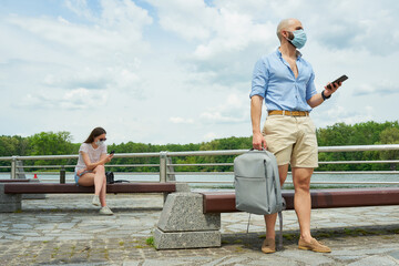 A bald man in a medical face mask keeping a social distance with a woman sitting on a bench on the promenade. A guy and a girl using smartphones in social distancing near the river in the afternoon.