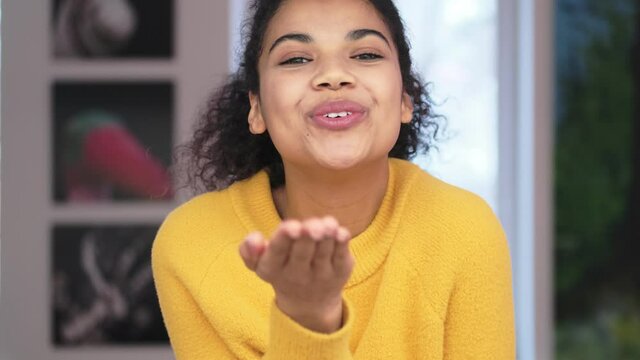 Cheerful african attractive woman in yellow sweater sending air kiss at the camera in living room